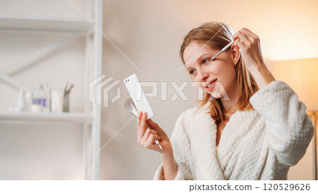 Beauty care. Female routine. Pleasant smiling lady applying makeup using powder brush looking in mirror enjoying her reflection in white robe while holding cosmetic tools in hand. 120529626