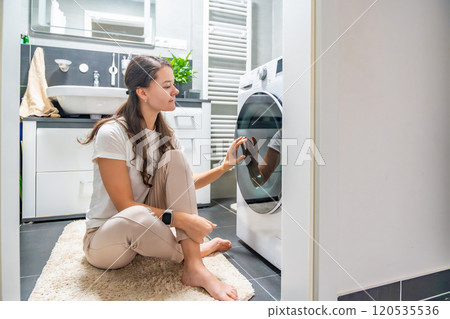 Young woman waiting for the washing machine to finish, looking at the washing machine door Young woman waiting for the washing machine to finish, looking at the washing machine door 120535536