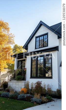 A modern white house with black window trim and a lush garden, set against a clear autumn sky 120538079