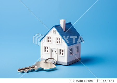 A miniature white house with a blue roof and a key placed beside it on a plain blue background 120538324