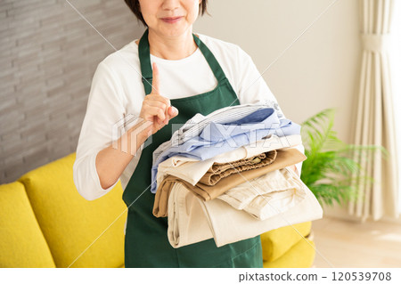A middle-aged to senior female housewife wearing a green apron holding laundry in the living room at home and pointing her index finger up 120539708