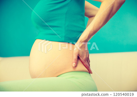 Pregnant woman sitting on couch holds hand on her back Pregnant woman sitting on couch holds hand on her back 120540019
