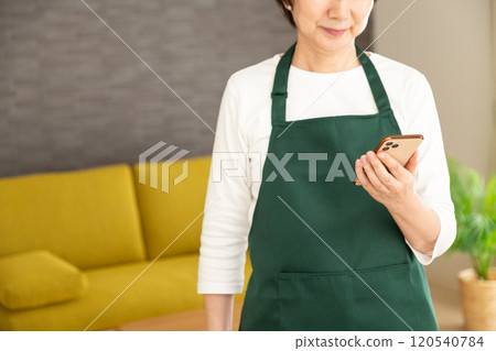 身穿綠色圍裙的中高級女家庭主婦在自家客廳看著智慧型手機 身穿綠色圍裙的中高級女家庭主婦在自家客廳看著智慧型手機 120540784
