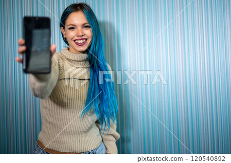 Portrait of a beautiful blue-haired hipster woman receiving a notification that causes her to have an extremely positive and surprised reaction. She extends her arm with her mobile phone towards us. 120540892