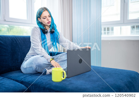 Beautiful blue haired hipster woman is working from home sitting on her couch, wearing headphones with microphone to attend a meeting with laptop. Concept of working from home, using technology. Beautiful blue haired hipster woman is working from home sitting on her couch, wearing headphones with microphone to attend a meeting with laptop. Concept of working from home, using technology. 120540902