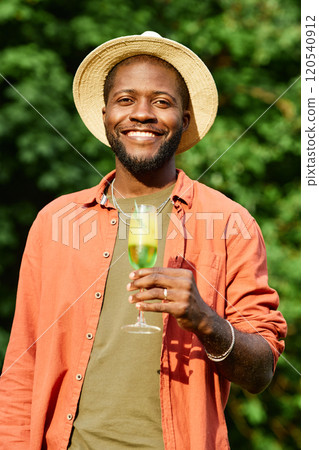 Smiling African American man wearing a hat and necklace while holding a drink and standing outdoors in natural surroundings, exuding a joyful and relaxed vibe 120540912
