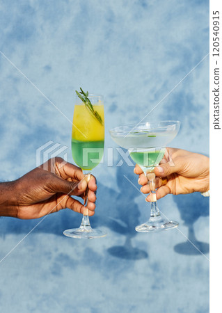 Close-up of two hands holding colorful cocktails against a blue background with a refreshing and celebratory vibe evident in the image 120540915