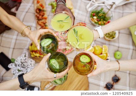 Friends raising glasses in toast at outdoor picnic, enjoying vibrant spread of food and drinks. Diverse hands and bracelets add unique charm to this festive gathering Friends raising glasses in toast at outdoor picnic, enjoying vibrant spread of food and drinks. Diverse hands and bracelets add unique charm to this festive gathering 120540925