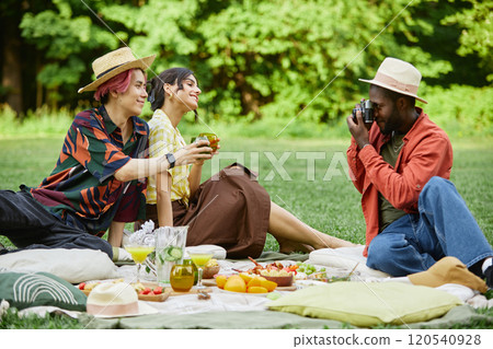 Group of friends sitting on blanket in green park having picnic with assortment of food, while one taking photo with camera, creating memorable moments 120540928
