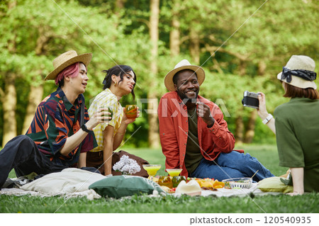 Group of friends sitting on picnic blanket in lush green park, sharing drinks and snacks, one person capturing memories with smartphone while others engage in cheerful conversation Group of friends sitting on picnic blanket in lush green park, sharing drinks and snacks, one person capturing memories with smartphone while others engage in cheerful conversation 120540935