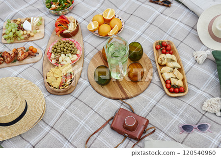 Array of vibrant picnic food displayed on checkered blanket outdoors including fruits, olives, salads, beverages in glasses, and accessories like hats and sunglasses arranged nearby 120540960
