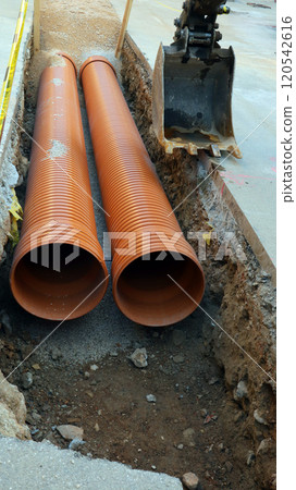 An excavator bucket is seen alongside two orange utility pipes that are laid in a trench. The excavation work reveals earthy surroundings and construction activity in progress. 120542616