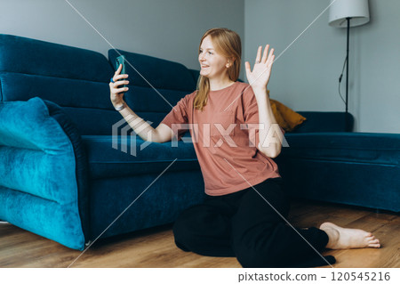 Hi. Female taking a selfie photo using her mobile phone at home. woman in t-shirt recording video, talking on online call and relaxing on floor near sofa. Communication and comfort concept, full body 120545216