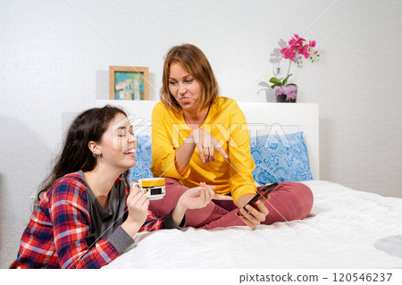 Two young Caucasian women are relaxing in the bedroom, chatting and looking at something on their smartphone with a smile. Concept of LGBT relations and social networks Two young Caucasian women are relaxing in the bedroom, chatting and looking at something on their smartphone with a smile. Concept of LGBT relations and social networks 120546237