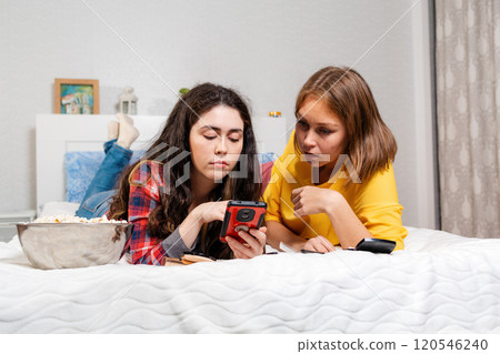 Two young Caucasian women are lying on a bed and looking at something on a smartphone, a bowl of popcorn is lying next to them. The concept of relationships , LGBT and communication Two young Caucasian women are lying on a bed and looking at something on a smartphone, a bowl of popcorn is lying next to them. The concept of relationships , LGBT and communication 120546240
