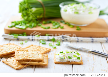 Crispy crackers and a glass bowl with a cream cheese spread and cut chives on a light kitchen table. 120552341