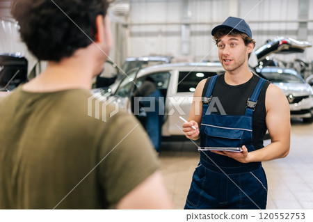 Male garage worker in uniform and unrecognizable man customer talking to discuss details of car repairs. Auto mechanic holding tablet and writes repair list for client in auto repair workshop. Male garage worker in uniform and unrecognizable man customer talking to discuss details of car repairs. Auto mechanic holding tablet and writes repair list for client in auto repair workshop. 120552753