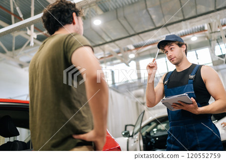 Low-angle view of male garage worker in uniform and unrecognizable man customer talking to discuss details of car repairs. Auto mechanic holding tablet and writes repair list for client Low-angle view of male garage worker in uniform and unrecognizable man customer talking to discuss details of car repairs. Auto mechanic holding tablet and writes repair list for client 120552759