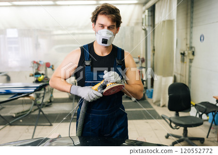 Portrait of professional car service male worker polishing black luxury car hood in auto repair shop using with orbital polisher, looking at camera. Concept of car detailing and polishing. 120552772