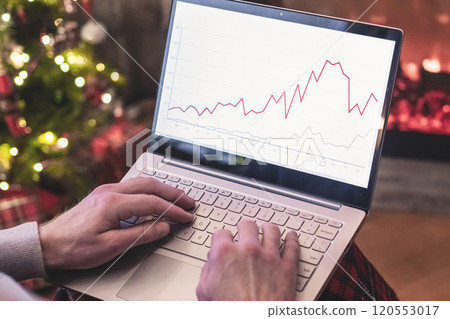 Man freelancer in santa claus hat working on laptop with graphs and charts on screen sitting near christmas tree and fireplace. 120553017