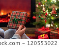 Woman in headphones with christmas gift box in hand sitting on fluffy plaid near fireplace and christmas tree. 120553022