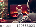 Woman in headphones with cup of hot cocoa and marshmallow sitting and warming at winter evening near fireplace flame and  christmas tree. 120553023