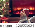 Woman with cup of hot cocoa and marshmallow sitting and warming at winter evening near fireplace flame and  christmas tree. 120553024