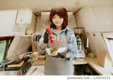 Asian relaxed female cook adding handmade ketchup on fresh grilled hot dog while standing in kitchen of food truck Asian relaxed female cook adding handmade ketchup on fresh grilled hot dog while standing in kitchen of food truck 120554375