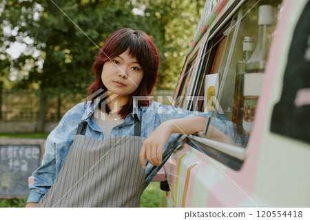 Medium close up of Asian beautiful female food truck worker in stripped apron leaning on pastel colored van 120554418