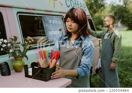 Medium close up of Asian young woman with stripped apron adjusting plastic box with multicolored straws and paper tissues on table next to food truck Medium close up of Asian young woman with stripped apron adjusting plastic box with multicolored straws and paper tissues on table next to food truck 120554464