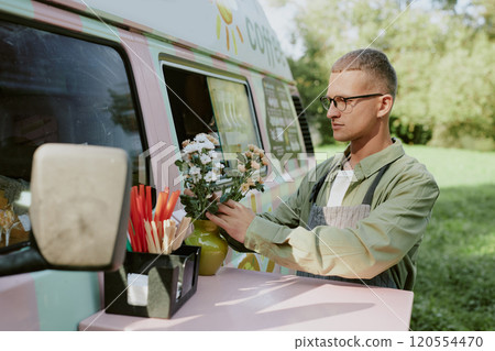 Young adult Caucasian man adjusting wildflowers in green clay vase on table next to food truck Young adult Caucasian man adjusting wildflowers in green clay vase on table next to food truck 120554470