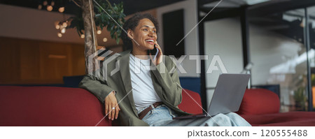 Woman in formal wear sitting on couch, talking on cell phone, using laptop in nice modern coworking office 120555488