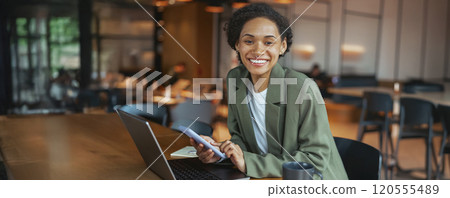 A woman in a suit smiles at her laptop and phone on the table A woman in a suit smiles at her laptop and phone on the table 120555489