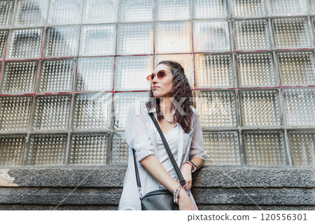Young woman in stylish sunglasses poses against a glass block wall 120556301