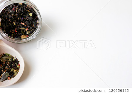 Large-grain tea with the addition of pieces of fruit and berries lies on a wooden spoon, next to it is a tilted glass jar with tea on a white background with space for text, Concept of tea drinking 120556325