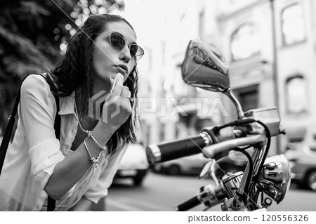Woman applying lipstick near motorcycle on city street in bright daylight Woman applying lipstick near motorcycle on city street in bright daylight 120556326