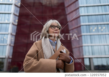 An elderly woman with glasses poses confidently in front of a modern building with glass and red An elderly woman with glasses poses confidently in front of a modern building with glass and red 120557052