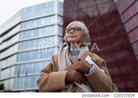 Older woman holding a laptop and standing confidently in front of modern glass buildings on a cloudy Older woman holding a laptop and standing confidently in front of modern glass buildings on a cloudy 120557054