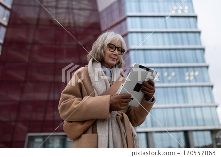 An elderly woman in a brown coat using a tablet outdoors in front of modern buildings on a cloudy 120557063