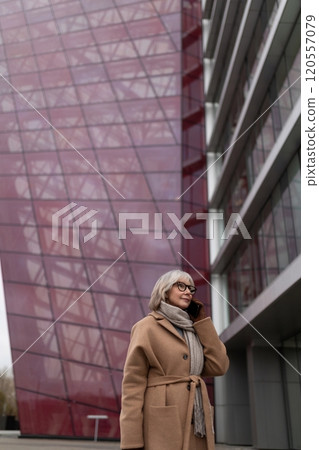A woman in a beige coat and scarf talks on her phone beside a modern building with a striking purple 120557079