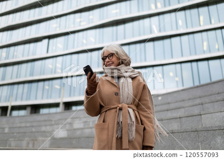 Elderly woman enjoying a moment while checking her phone near a modern building in a bustling urban 120557093