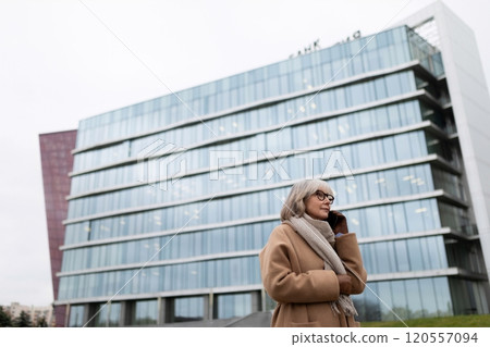 Elderly woman in brown coat talks on mobile phone outside modern glass office building in a city Elderly woman in brown coat talks on mobile phone outside modern glass office building in a city 120557094