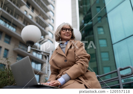 Woman working on laptop outside a modern building in an urban setting during daylight hours Woman working on laptop outside a modern building in an urban setting during daylight hours 120557118