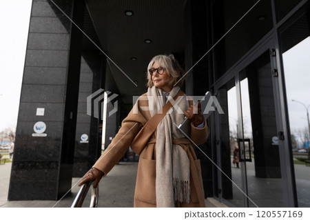 Senior woman in stylish coat holding tablet exits modern building on a chilly day 120557169