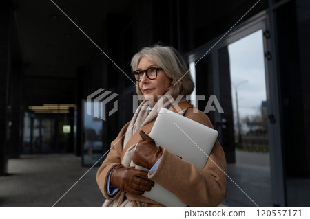 An elegant woman stands outside a modern building holding a laptop, wearing a warm coat and glasses An elegant woman stands outside a modern building holding a laptop, wearing a warm coat and glasses 120557171