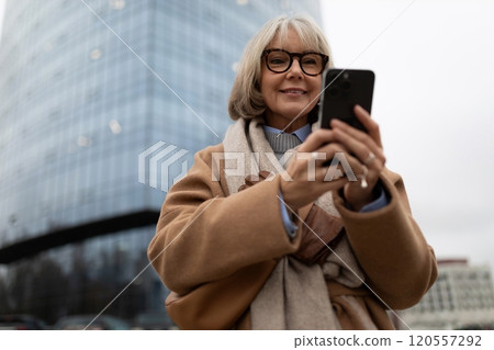 Older woman smiling while using smartphone in urban setting with modern buildings around her during 120557292