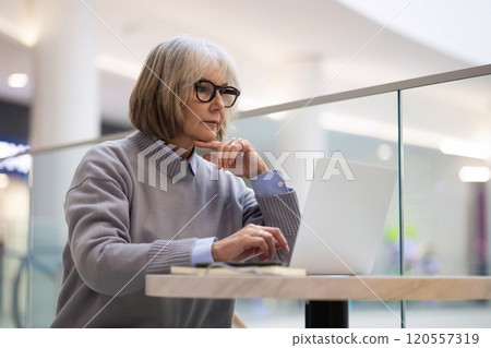 Senior woman working on laptop at a cafe in a modern shopping mall during the daytime, focused on 120557319
