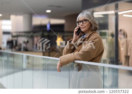 A mature woman wearing glasses talks on her phone while leaning against a railing in a modern A mature woman wearing glasses talks on her phone while leaning against a railing in a modern 120557322