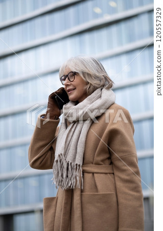 A smiling elderly woman in a brown coat talks on her smartphone while standing outside a modern A smiling elderly woman in a brown coat talks on her smartphone while standing outside a modern 120557329
