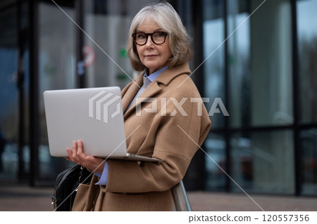 Senior woman with laptop outside modern building during daytime, showcasing confidence and 120557356